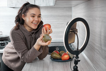 A young woman smiling in the kitchen with a ring lamp and a phone shares healthy food recipes. The concept of Food blogger.