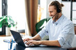 Satisfied modern male manager or call center worker in headset is looks at the laptop screen, having online business meeting. Successful employee talking online with colleagues uses laptop