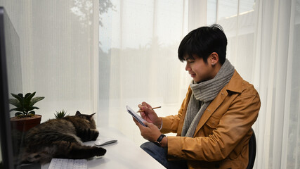 Wall Mural - Side view of young man designer working online at home with digital tablet and cute cat lying in front of him.