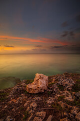 Wall Mural - Sunset view. Seascape background. Amazing ocean view. Beach with rocks and stones. Bright sunlight on horizon line. Cloudy sky. Vertical layout. Bali.