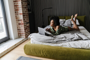 Wall Mural - Focused african american girl using laptop while lying in bed after sleep