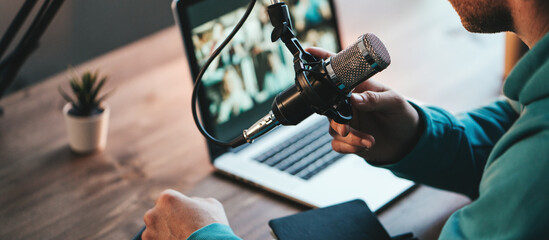 A man host streaming his audio podcast using microphone and laptop at his small broadcast studio, close-up