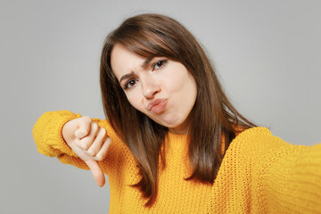 Wall Mural - Close up young smiling friendly positive attractive woman in casual knitted yellow sweater do selfie shot on mobile phone show thumb down dislike gesture isolated on grey background studio portrait.