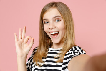 Wall Mural - Close up little blonde smiling happy kid girl 12-13 years old in striped oversized t-shirt do selfie shot on mobile phone show ok okay gesture isolated on pink background children Childhood concept.