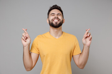 Wall Mural - Young caucasian bearded happy man 20s wear casual yellow basic t-shirt waiting for special moment, keeping fingers crossed, making wish, eyes closed, isolated on grey color background studio portrait.