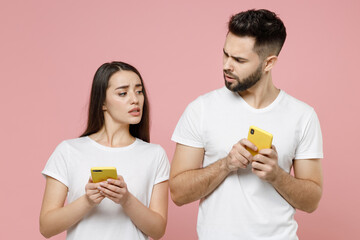 Poster - Young shocked couple two friends man woman in white basic blank print design t-shirts hold in hand using mobile cell phone pry into chatting typing sms spy on peep isolated on pastel pink background.