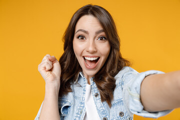 Wall Mural - Close up of young excited happy fun surprised woman in denim shirt white t-shirt do selfie shot on mobile phone do winner gesture clench fist celebrating isolated on yellow background studio portrait