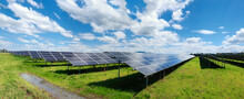 Solar Power Plant, Blue Solar Panels On Orange Autumn Grass Field Under Blue Sky With Clouds. Toned Panoramic Image. Solar Power Generation, Renewable Energy Production