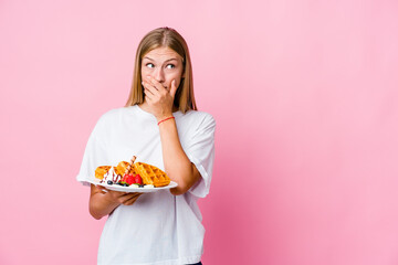 Wall Mural - Young russian woman eating a waffle isolated thoughtful looking to a copy space covering mouth with hand.