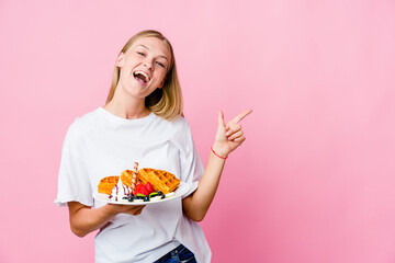 Wall Mural - Young russian woman eating a waffle isolated pointing with forefingers to a copy space, expressing excitement and desire.