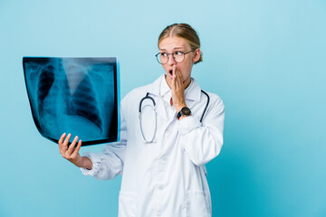 Young russian doctor woman holding a bone scan on blue is saying a secret hot braking news and looking aside