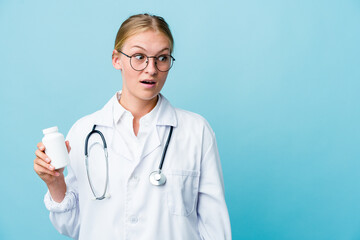 Wall Mural - Young russian doctor woman holding pills bottle on blue being shocked because of something she has seen.