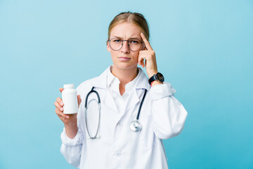 Wall Mural - Young russian doctor woman holding pills bottle on blue focused on a task, keeping forefingers pointing head.