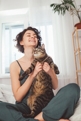 Wall Mural - Woman sitting at home on the bed and laughing while holding her cat