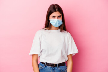 Young caucasian woman wearing a mask for virus isolated on pink background shrugs shoulders and open eyes confused.