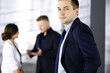 © cameravit - Portrait of a self-confident middle aged businessman in a blue suit, standing in a modern office with his colleagues at the background. Concept of business success
