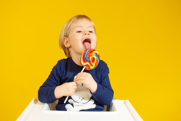 Cute little boy eating colorful lollipop while against sitting on chair over yellow background.