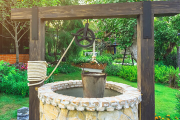 Beautiful artesian well made by stones and wheel pulley with metal bucket and rope in peaceful garden atmosphere. Retro stone water well in rural area. Garden decoration with antique items.