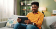 © VAKSMANV - Portrait of African American joyful happy young man resting on couch in living room in apartment and tapping on tablet device browsing online searching internet and looking away. Spare time concept