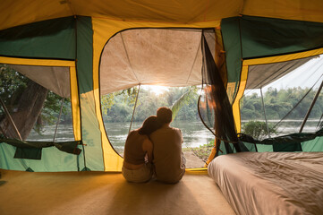 Wall Mural - Young asian couple embracing and relaxing inside a tent on campsite in natural park