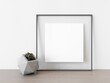 © Washdog - Empty grey square frame stands on light wood table against wall. Mockup of poster frame close up in home interior with succulent plant, 3D illustration