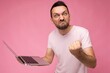 © Ivan Traimak - Handsome angry man holding laptop computer showing fist looking at camera in t-shirt on isolated pink background
