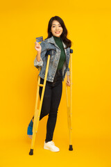 Portrait of a happy young Asian woman using a crutch and showing credit card on hand isolated on yellow background, Foot injury and Personal accident concept