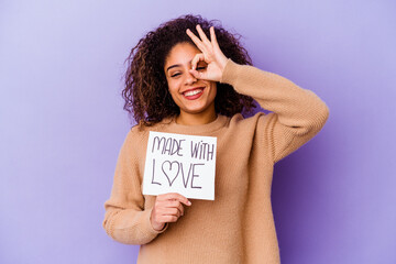 Young African American woman holding a Made with love placard isolated on purple background excited keeping ok gesture on eye.