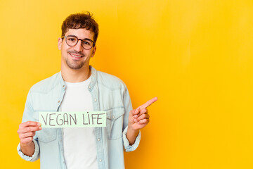 Wall Mural - Young caucasian man holding a vegan life placard isolated smiling and pointing aside, showing something at blank space.