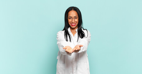 Wall Mural - young black woman smiling happily with friendly, confident, positive look, offering and showing an object or concept. physician concept