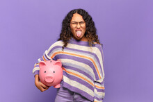 Young Hispanic Woman Feeling Disgusted And Irritated, Sticking Tongue Out, Disliking Something Nasty And Yucky. Piggy Bank Concept
