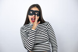 Fototapeta  - Young beautiful brunette burglar woman wearing mask hand on mouth telling secret rumor, whispering malicious talk conversation