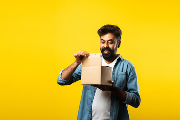 Indian Handsome bearded young man opening box with parcel while standing against yellow background