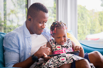 Wall Mural - Cheerful african american father and daughter playing in living room, Cute little girl and dad sitting on the sofa and playing on tablet, Happiness family concepts