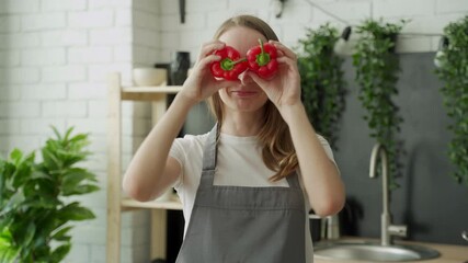 Wall Mural - Happy young woman smiles and has fun playing with red pepper in front of her eyes in the home kitchen