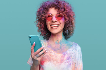 Happy curly haired woman with Holi colors using smartphone