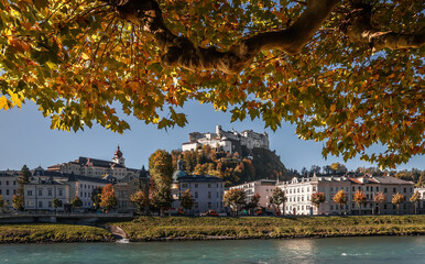 Fototapete - Sunny Autumnal scenic cityscape with historical buildings in Salzburg city. Wonderful view on  famous Hohensalzburg Fortress, Salzach river and colorful trees under sunlight.
