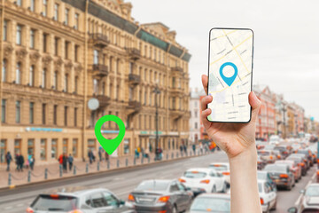 A female's hand holds a smartphone with an online map app. In the background is a blurred city street with traffic, with a green location icon.Concept of online navigation and GPS