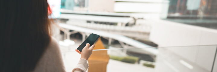 Shoulder view of woman using mobile phone empty screen with blur city banner size background.