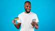 © Prostock-studio - Cheerful Black Guy Using Phone And Credit Card, Blue Background