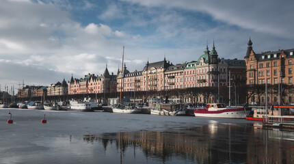 Wall Mural - City pier (Stockholm) in winter. Surface is partially frozen with partial reflection of scenery. Boats are moored and winterized. Trees on waterfront are fallen and sky is partially cloudy.