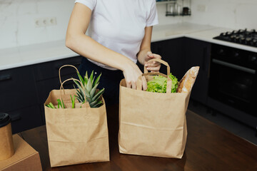 Home food delivery. Various vegetables and fruits in a paper bags that a young girl is sorting out at home on the kitchen table.