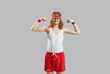 Funny thin skinny man with long hair in eyeglasses, retro headband, white tank top and red shorts showing weak muscles standing isolated on gray background. Sports workout and fitness exercise concept
