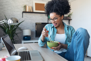 Wall Mural - Beautiful young afro business woman making video call with laptop while eating noodles with chopsticks sitting in the office at home.