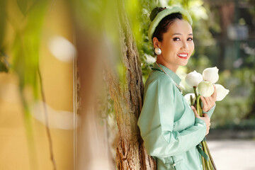 Poster - Portrait of young happy Vietnamese woman in ao dai dress and traditional headser holding lotus flowers when standing in city park