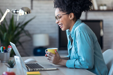 Beautiful young afro business woman making video call with laptop while drinking coffee sitting in the office at home.