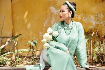 Poster - Attractive Vietnamese young woman in mint green ao dai dress and traditional headwear sitting outdoors with lotus flowers in hands