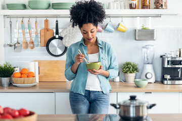 Wall Mural - Beautiful afro woman eating noodles with chopsticks while standing in the kitchen at home.