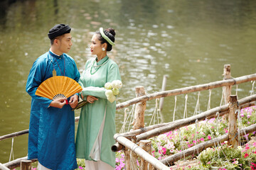 Poster - Just married Vietnamese husband and wife in traditional dresses standing on wooden bridge and smiling at each other