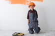 © Andrii Lysenko - a little boy dressed in a large overalls and an orange helmet on his head is standing near the wall.